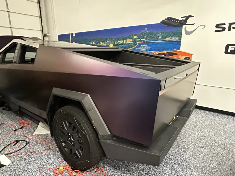 Rear quarter of a Cybertruck in a satin color-shift film, reading purple across the bed side and near-black in shadow
