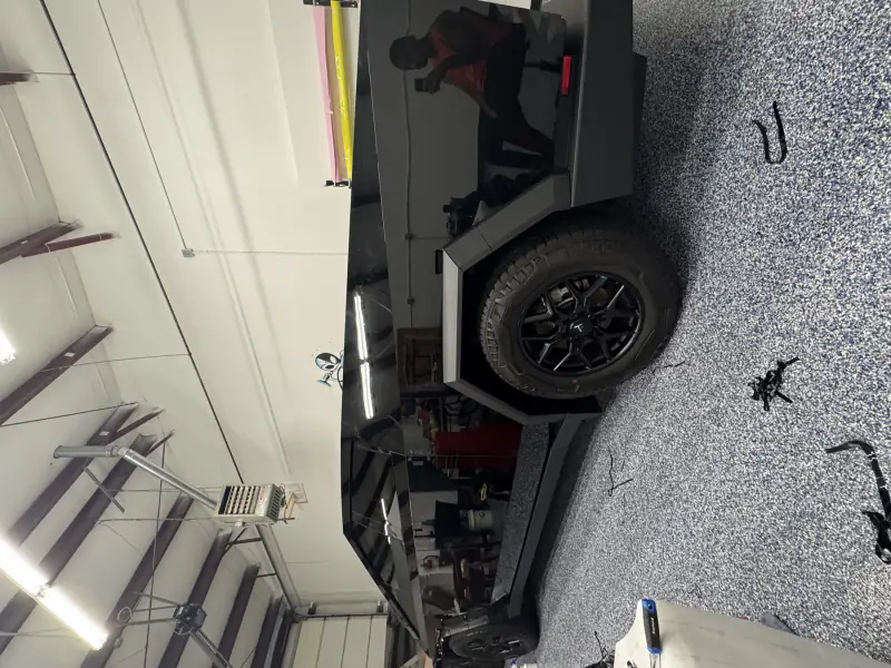 Cybertruck wrapped in gloss black under the bay lights, the panels reflecting the shop