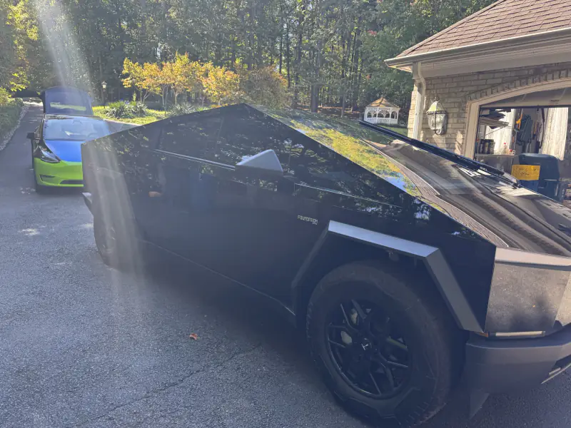 Cybertruck in gloss black on a residential driveway, a wrapped Tesla behind it