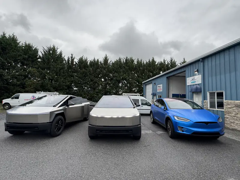 Two Cybertrucks in bare stainless beside a Model X wrapped in satin blue, outside the bay