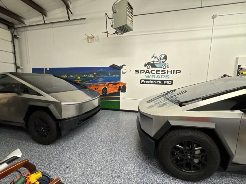 Two Cybertrucks in the Frederick, Maryland bay, beside the Tesla Wrapper wall logo