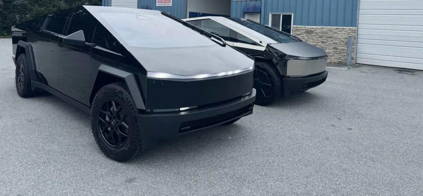 A Cybertruck finished in gloss black parked beside an unwrapped one in bare stainless, both in the same daylight