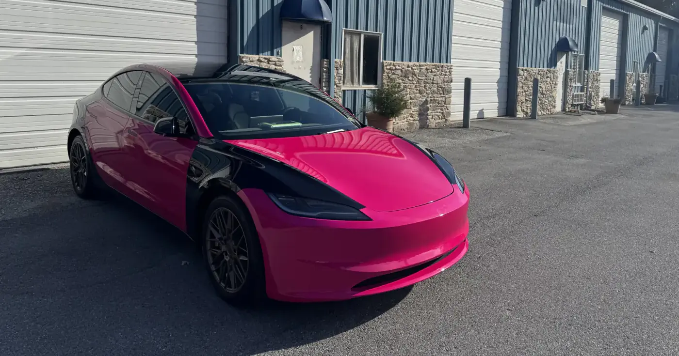 A Tesla Model 3 wrapped in hot pink with a black hood flash, photographed in sunlight outside the shop