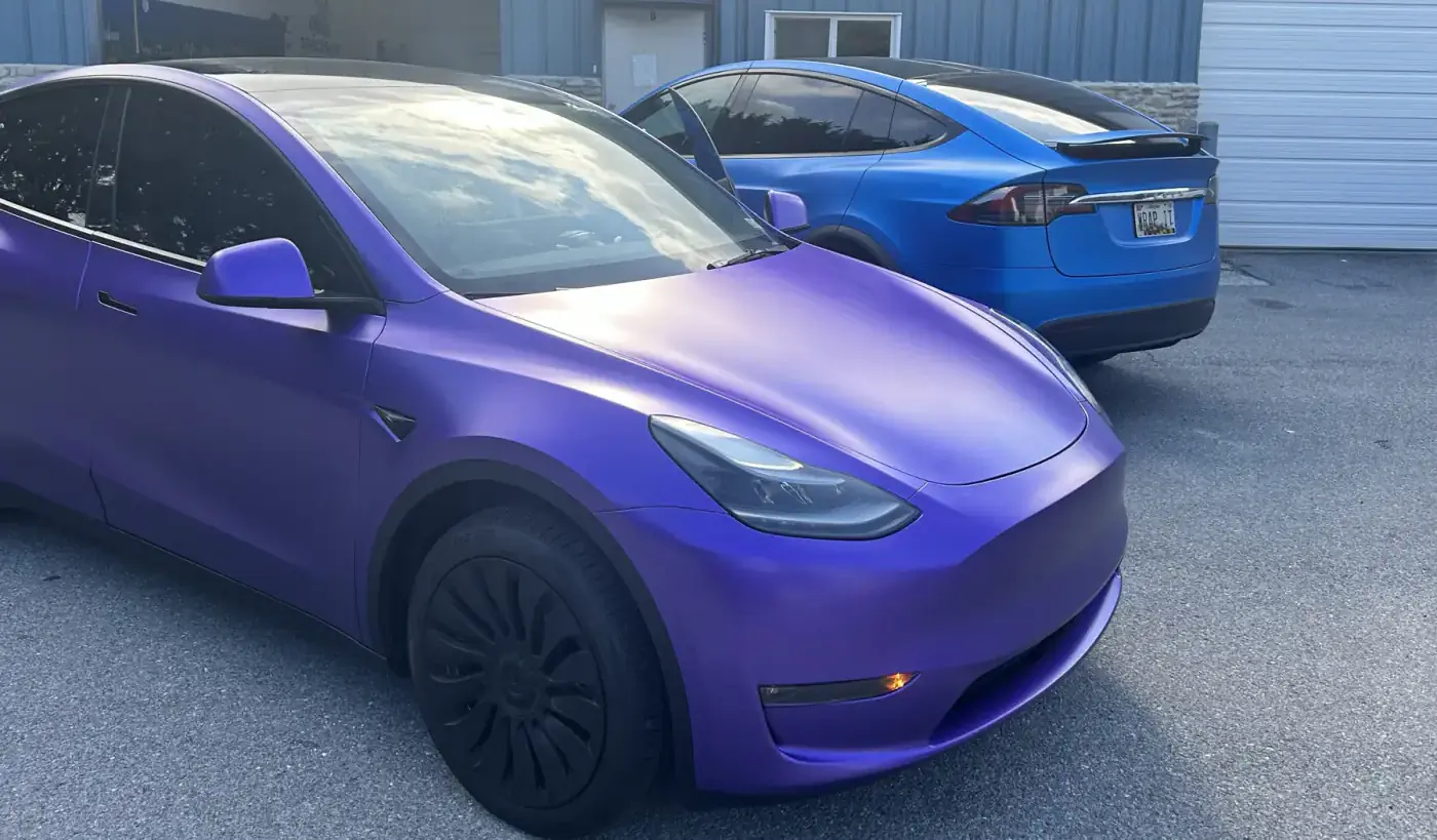 A Tesla Model Y wrapped in satin purple in evening light, with a wrapped Model X behind it