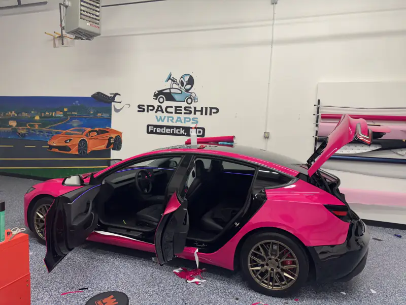A hot pink Model 3 with doors and trunk open mid-install, offcuts of film on the floor