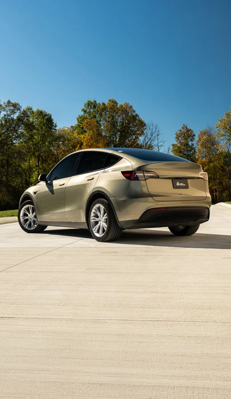 The rear three-quarter of a Tesla Model Y in PrismaPro Midnight Sand, parked outdoors in sunlight